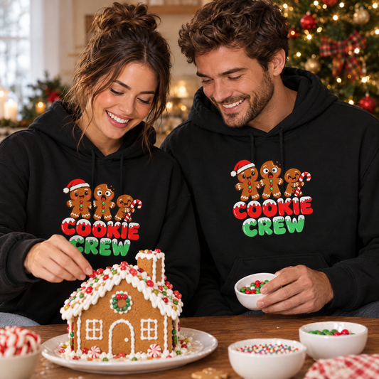 Couple in matching black Cookie Crew hoodies decorating a gingerbread house by the Christmas tree